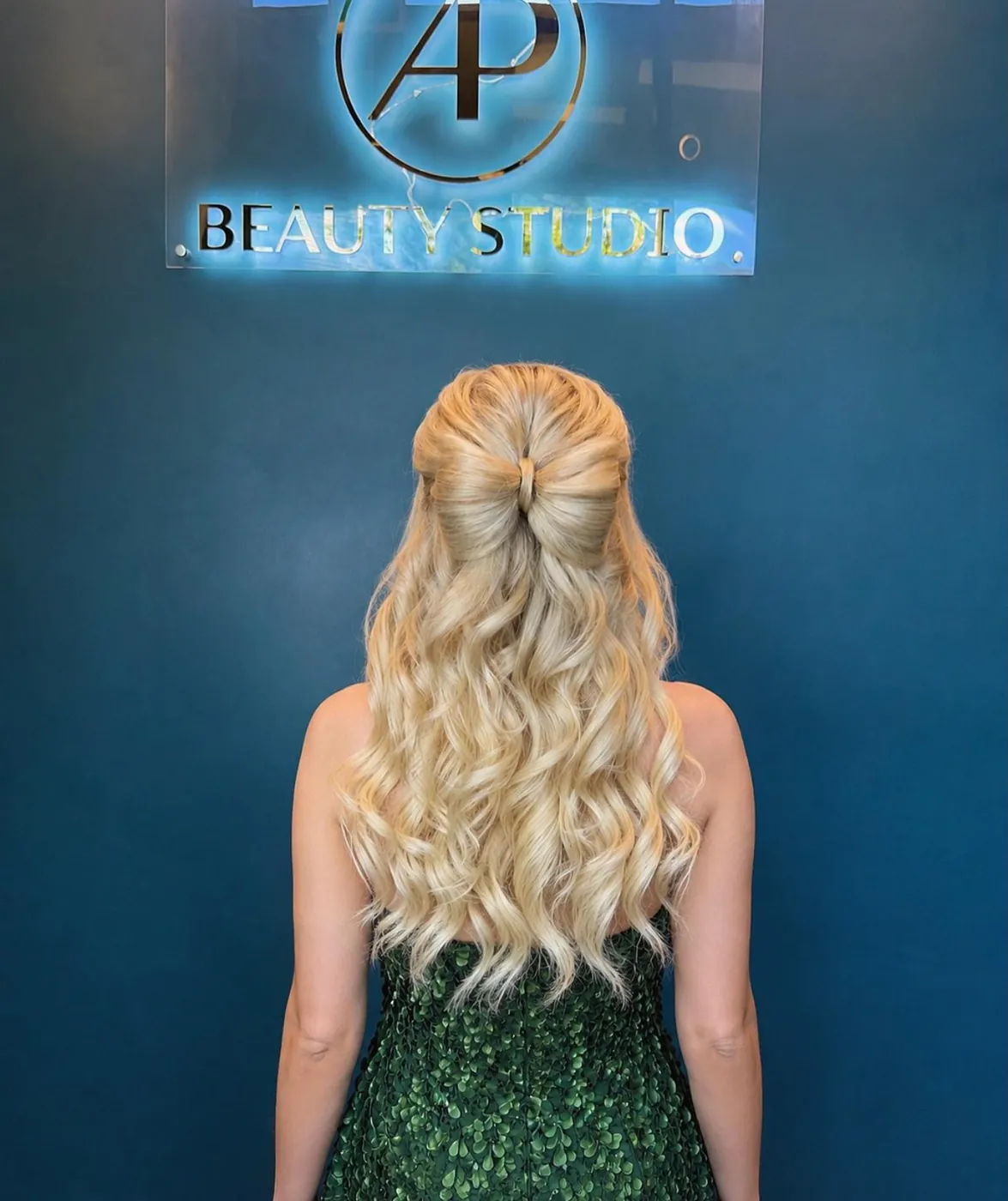 Blonde curls styled half up with a bow, in front of the AP Beauty Studio sign