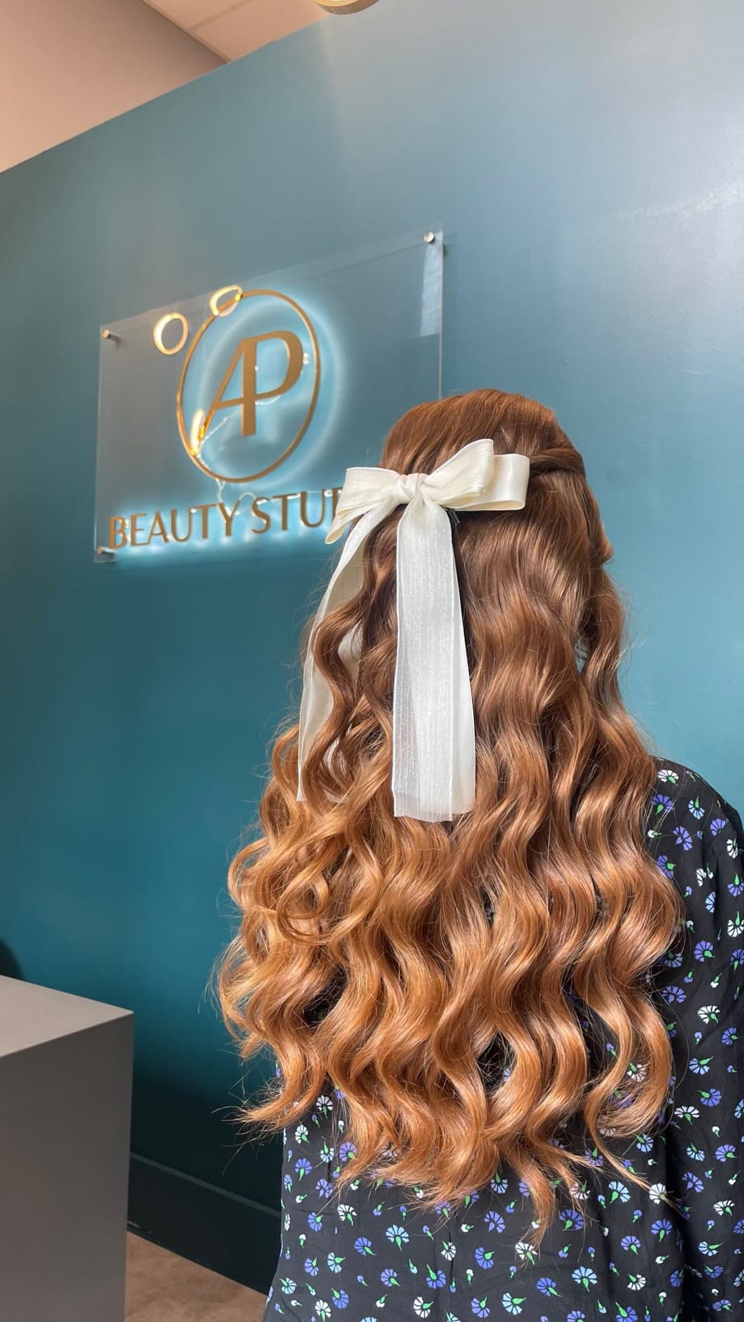 Honey-copper curls with a white bow, in front of the AP Beauty Studio sign